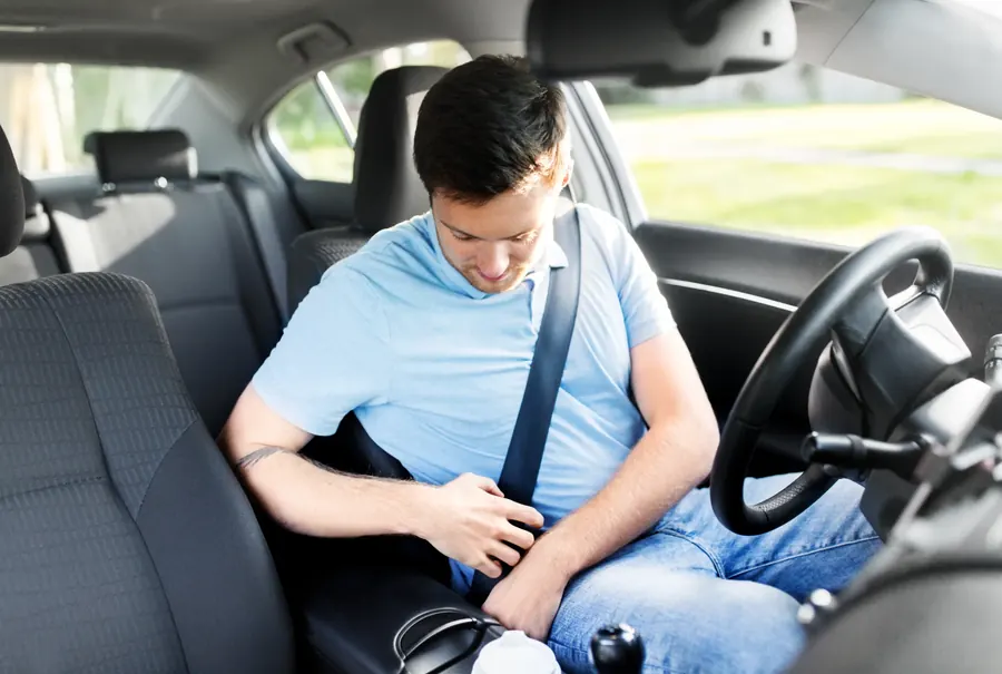 A seated driver fastening the seatbelt buckle inside a car with the steering wheel and rear seats visible.