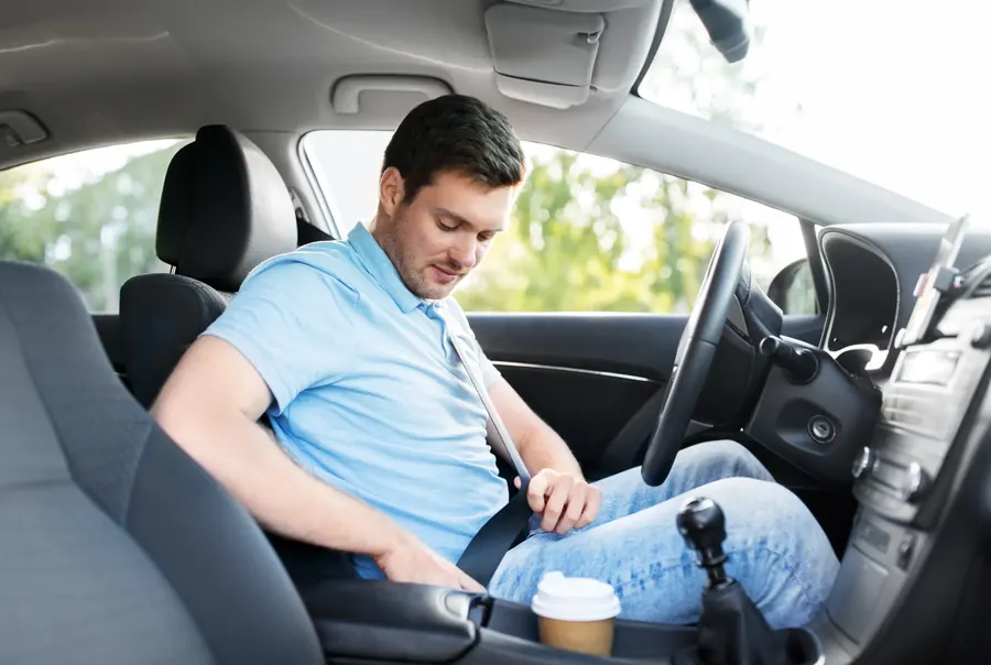 A driver seated in a car looking down as he fastens the seatbelt across his chest and hip.