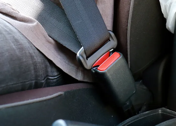 A fastened seatbelt across a seated occupant&rsquo;s hip, with the latch plate engaged in a red-release buckle beside the seat.