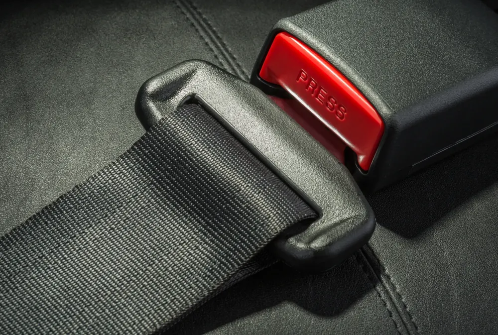 Black seatbelt webbing threaded through a moulded latch plate beside a red-release buckle housing on a dark leather seat.