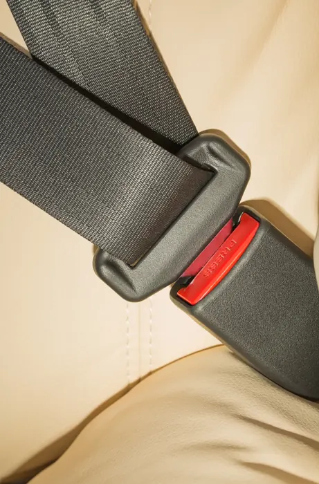 A dark seatbelt latch plate engaged in a buckle with a red release button against a cream leather seat.