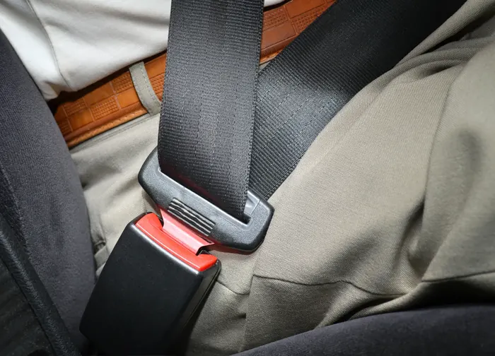 A fastened black seatbelt crossing a seated person&rsquo;s lap, with the latch plate engaged in a red-release buckle.